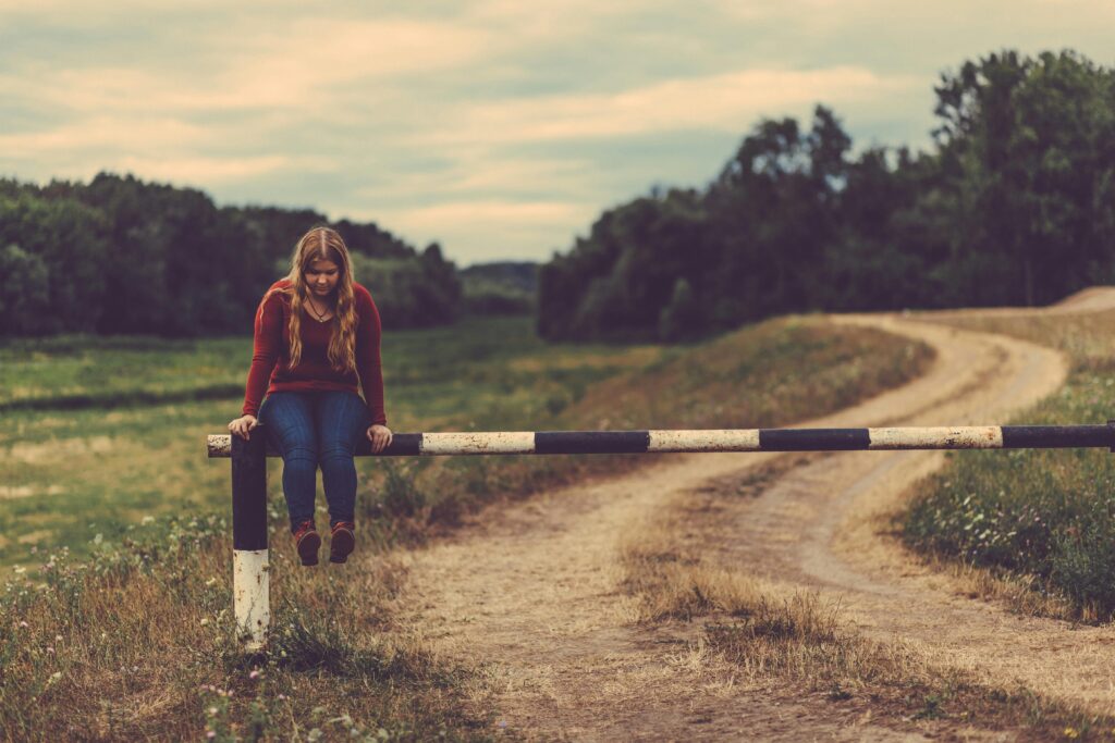 A young woman sits on a barrier in a scenic rural path surrounded by fields and trees.